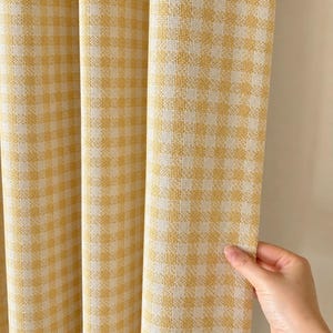 Può includere: Un pannello per tenda caratterizzato da un classico motivo a quadretti vichy gialli e bianchi. Il tessuto presenta una trama visibile con pieghe verticali che creano profondità. Il materiale è opaco e mostra un disegno a griglia uniforme.