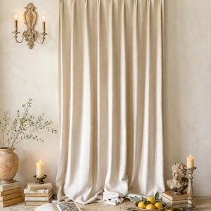 May include: A floor-length, off-white curtain with a pinch-pleat header hangs from a wooden rod. The fabric features a subtle, textured weave and falls in vertical folds, reaching the floor with a slight gather at the bottom edge.