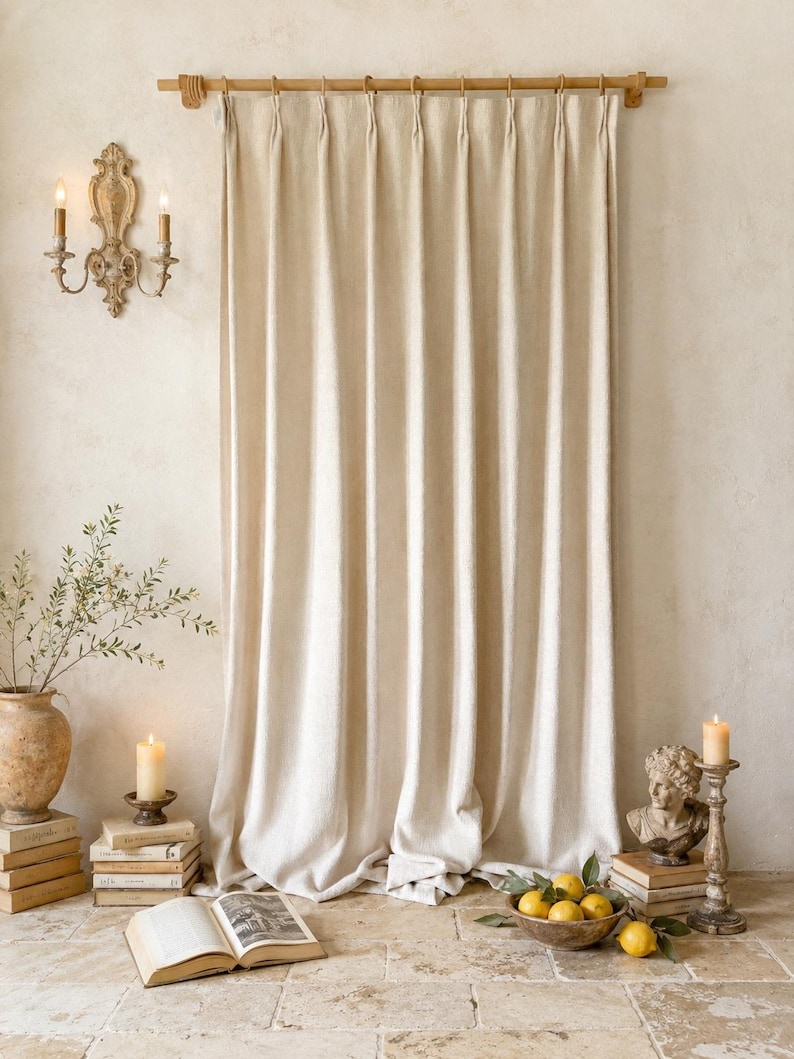May include: A floor-length, off-white curtain with a pinch-pleat header hangs from a wooden rod. The fabric features a subtle, textured weave and falls in vertical folds, reaching the floor with a slight gather at the bottom edge.