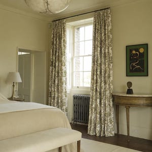 May include: A bedroom interior featuring a large bed with a beige coverlet and bench, a hanging alabaster bowl pendant light, floor-length curtains with a green and white botanical print, a dark gray radiator, and a semi-circular wooden console table.