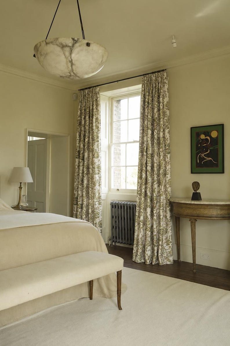May include: A bedroom interior featuring a large bed with a beige coverlet and bench, a hanging alabaster bowl pendant light, floor-length curtains with a green and white botanical print, a dark gray radiator, and a semi-circular wooden console table.