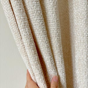 May include: A cream-colored curtain panel featuring a thick, textured, woven fabric with a soft, plush pile. The material displays a dense, grid-like pattern created by the interlacing of light-colored threads, resulting in a dimensional surface.