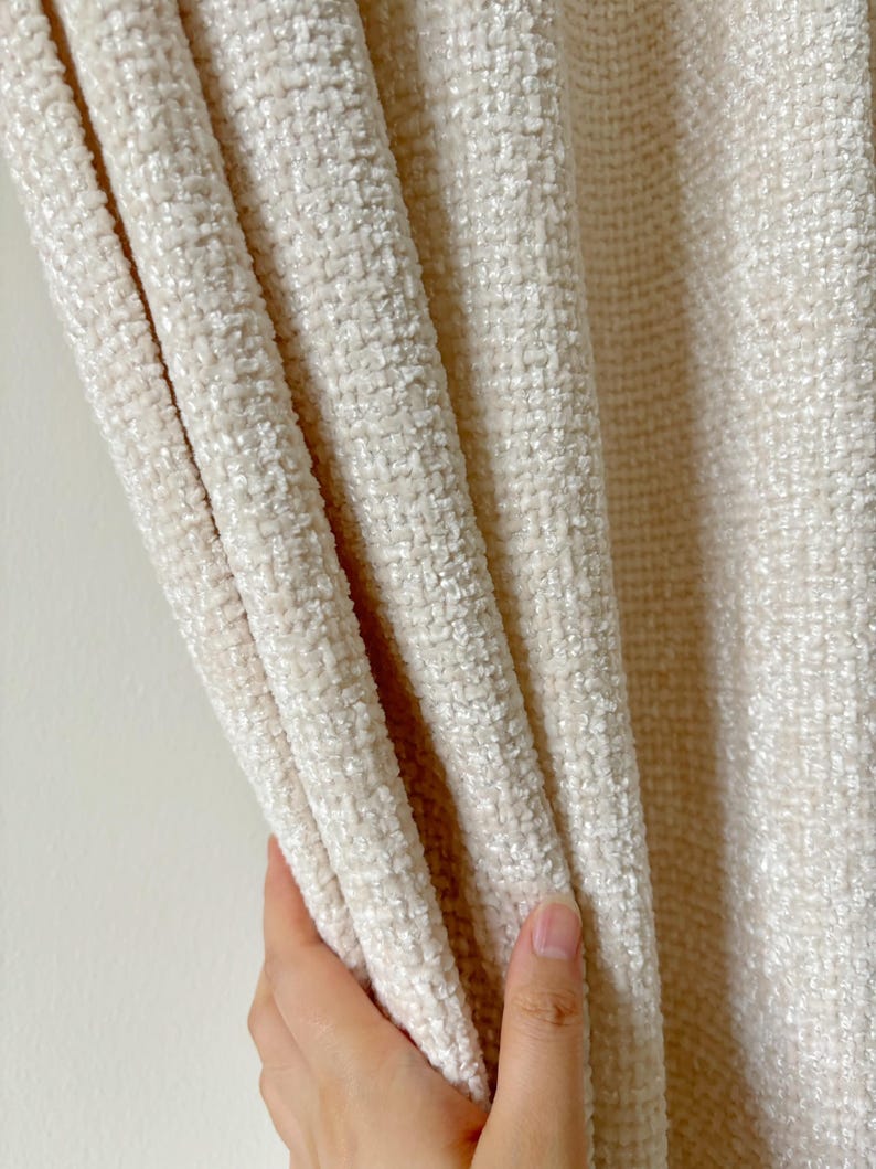 May include: A cream-colored curtain panel featuring a thick, textured, woven fabric with a soft, plush pile. The material displays a dense, grid-like pattern created by the interlacing of light-colored threads, resulting in a dimensional surface.
