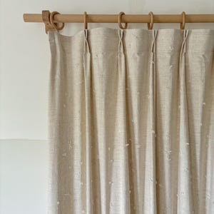 May include: Beige linen curtains hanging from a wooden rod with wooden rings. The curtains are pleated and have a subtle textured pattern. The rod and rings are a light wood color, creating a neutral and natural aesthetic.