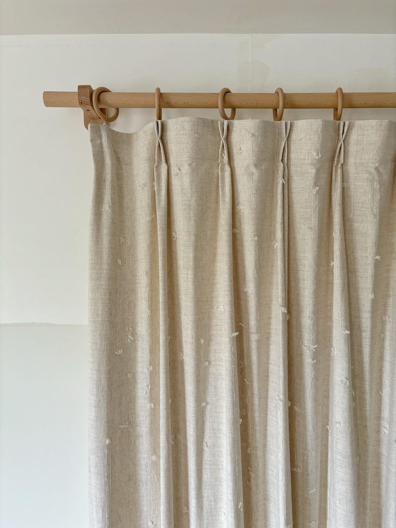 May include: Beige linen curtains hanging from a wooden rod with wooden rings. The curtains are pleated and have a subtle textured pattern. The rod and rings are a light wood color, creating a neutral and natural aesthetic.