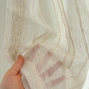 May include: A sheer, off-white fabric curtain featuring a subtle vertical striped pattern with light tan, textured fibers woven throughout. The material is lightweight and semi-transparent, allowing for a soft diffusion of light through the weave.