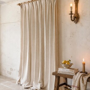 May include: A floor-length, off-white pleated curtain hangs from a wooden rod with rings. Beside it, a wooden bench holds a bowl of lemons, two stacked books titled "Couture Finishes" and "Maison de Fleur," a candle, and a ceramic pitcher.