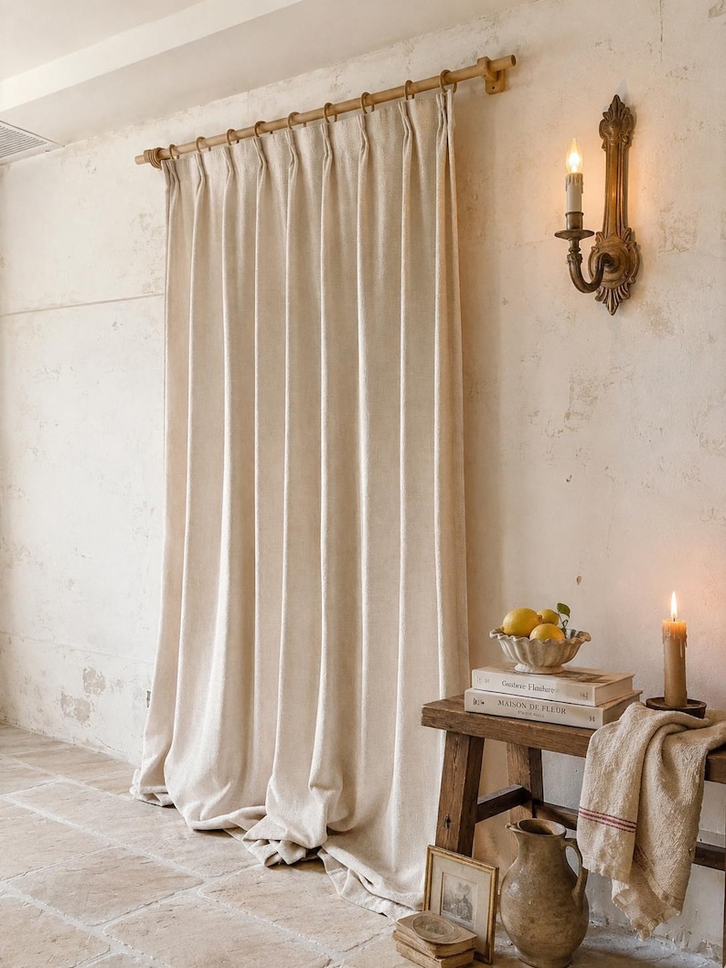 May include: A floor-length, off-white pleated curtain hangs from a wooden rod with rings. Beside it, a wooden bench holds a bowl of lemons, two stacked books titled "Couture Finishes" and "Maison de Fleur," a candle, and a ceramic pitcher.
