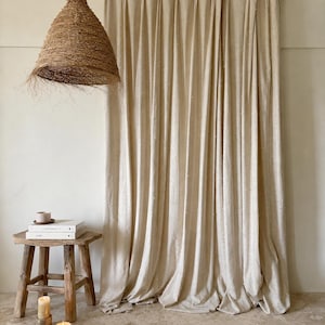 May include: A neutral-toned interior scene featuring a woven pendant light, a wooden stool with books and a cup, and a set of beige curtains. The curtains are hung on a wooden rod, and the floor is a light stone color. Candles are lit on the stool.