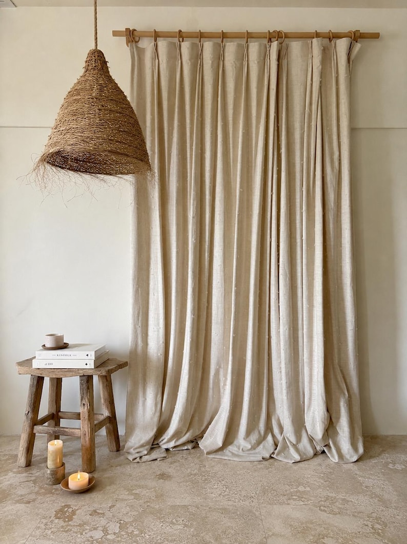 May include: A neutral-toned interior scene featuring a woven pendant light, a wooden stool with books and a cup, and a set of beige curtains. The curtains are hung on a wooden rod, and the floor is a light stone color. Candles are lit on the stool.