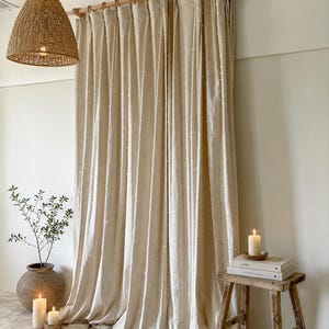May include: Beige linen curtains with a subtle dotted pattern hang from a wooden rod. A woven pendant light hangs from the ceiling. A small wooden stool holds books and a lit candle. A potted plant and more candles are on the stone floor.