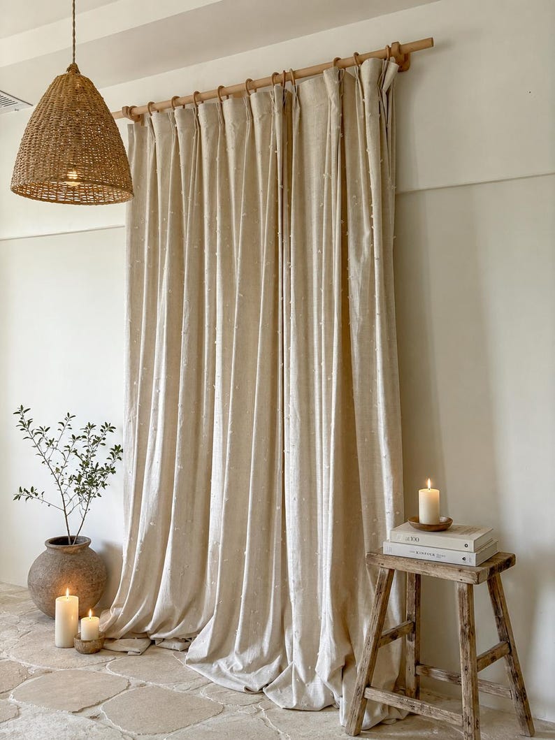 May include: Beige linen curtains with a subtle dotted pattern hang from a wooden rod. A woven pendant light hangs from the ceiling. A small wooden stool holds books and a lit candle. A potted plant and more candles are on the stone floor.