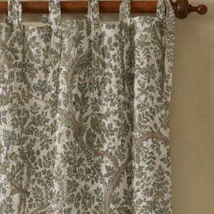 May include: A curtain panel featuring a detailed botanical print of brown tree branches and green leaves on a light beige fabric. The curtain is suspended from a dark wooden rod by several fabric tie-top loops along the upper edge.