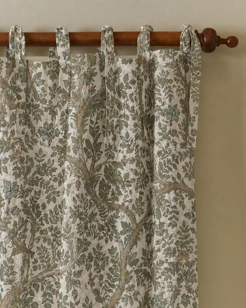 May include: A curtain panel featuring a detailed botanical print of brown tree branches and green leaves on a light beige fabric. The curtain is suspended from a dark wooden rod by several fabric tie-top loops along the upper edge.