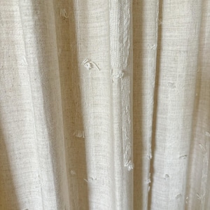 May include: Close-up of off-white fabric curtains with vertical textured stripes and frayed details. The curtains have a linen-like appearance, with a soft, neutral color palette. The fabric drapes in gentle folds, creating a sense of texture and depth.