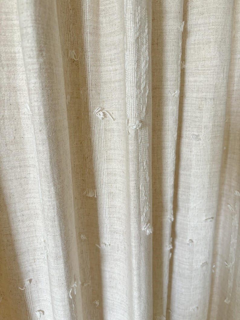 May include: Close-up of off-white fabric curtains with vertical textured stripes and frayed details. The curtains have a linen-like appearance, with a soft, neutral color palette. The fabric drapes in gentle folds, creating a sense of texture and depth.