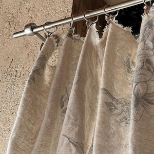 May include: Beige linen curtains with a grey floral pattern suspended from a silver rod. The curtains have a raw edge and are clipped to the rod with metal rings. The background is a textured, light brown wall.