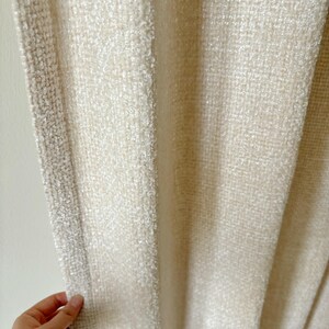 May include: A cream-colored curtain featuring a thick, textured weave with a prominent vertical border. The fabric displays a dense, nubby surface pattern that creates a consistent, uniform appearance across the entire length of the hanging panel.
