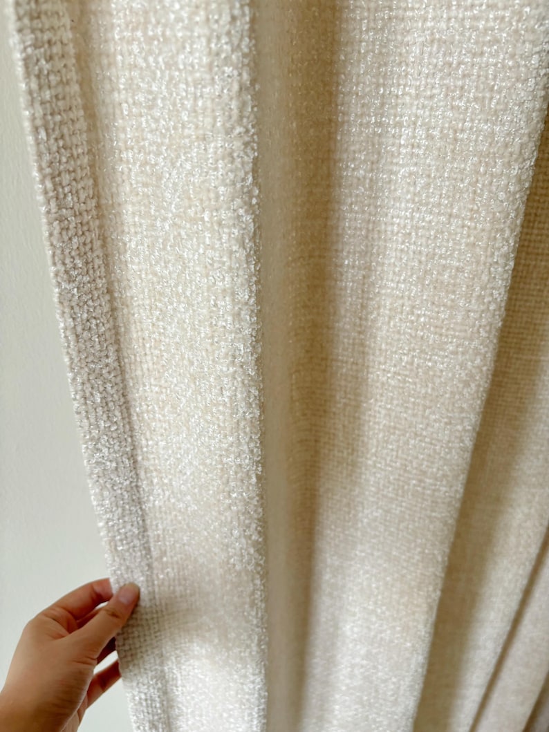 May include: A cream-colored curtain featuring a thick, textured weave with a prominent vertical border. The fabric displays a dense, nubby surface pattern that creates a consistent, uniform appearance across the entire length of the hanging panel.