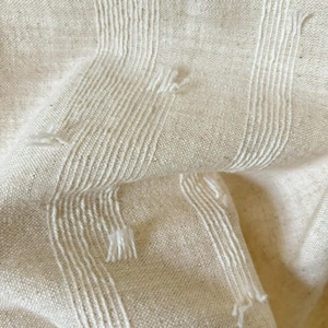 May include: Close-up of a textured, off-white fabric with vertical white stripes. The fabric has a woven appearance with visible threads and slight imperfections, creating a rustic aesthetic. The stripes have a fringed effect.