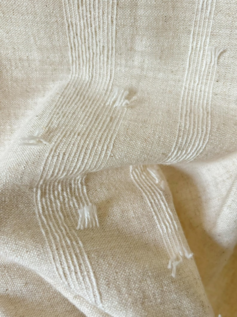 May include: Close-up of a textured, off-white fabric with vertical white stripes. The fabric has a woven appearance with visible threads and slight imperfections, creating a rustic aesthetic. The stripes have a fringed effect.