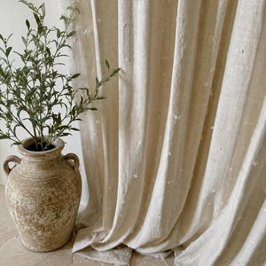 May include: Beige linen curtains with a textured, dotted pattern. The curtains are draped, revealing soft folds. A weathered, light brown ceramic vase with handles holds a green plant, adding a natural element to the scene.