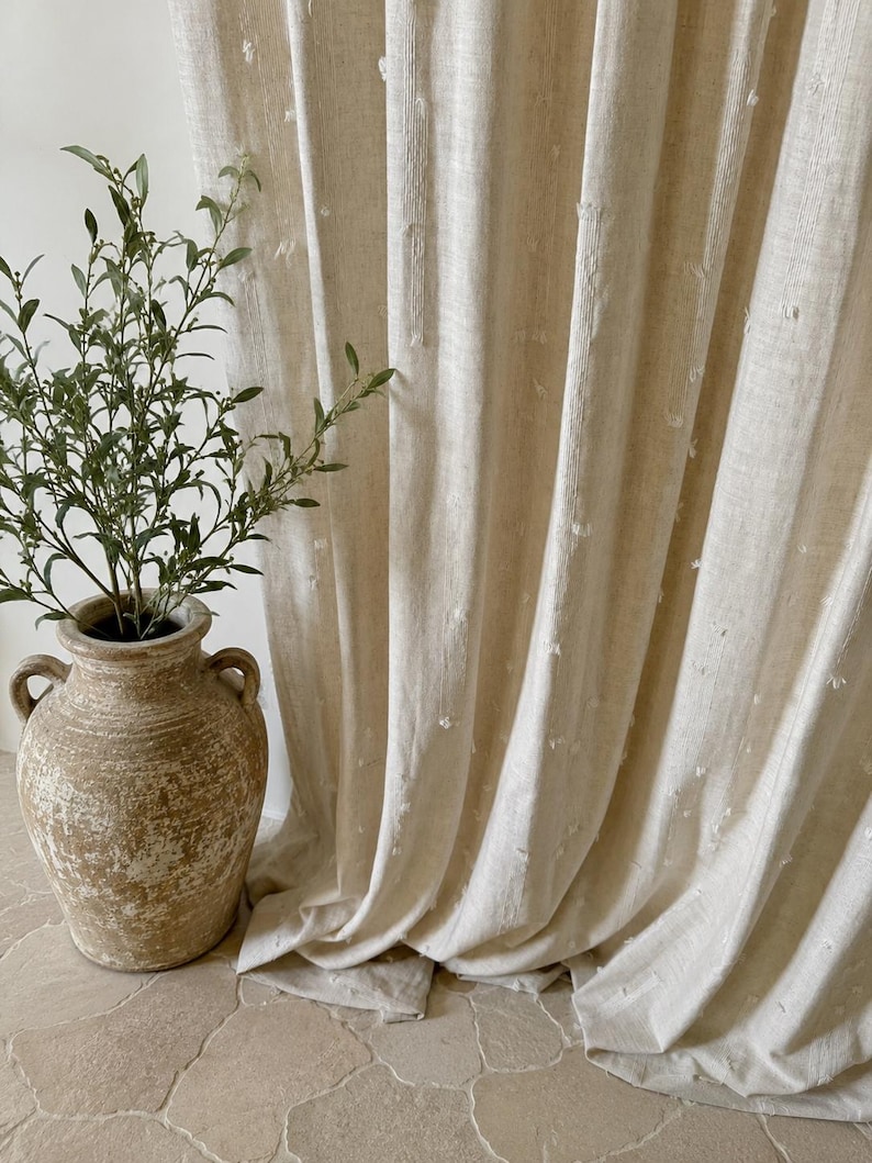 May include: Beige linen curtains with a textured, dotted pattern. The curtains are draped, revealing soft folds. A weathered, light brown ceramic vase with handles holds a green plant, adding a natural element to the scene.