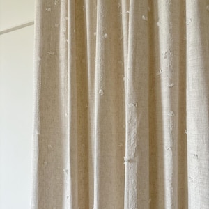 May include: Beige linen curtains with vertical folds. The fabric has a textured appearance with small, irregular white tufts scattered across the surface. The curtains are hanging in front of a white wall.