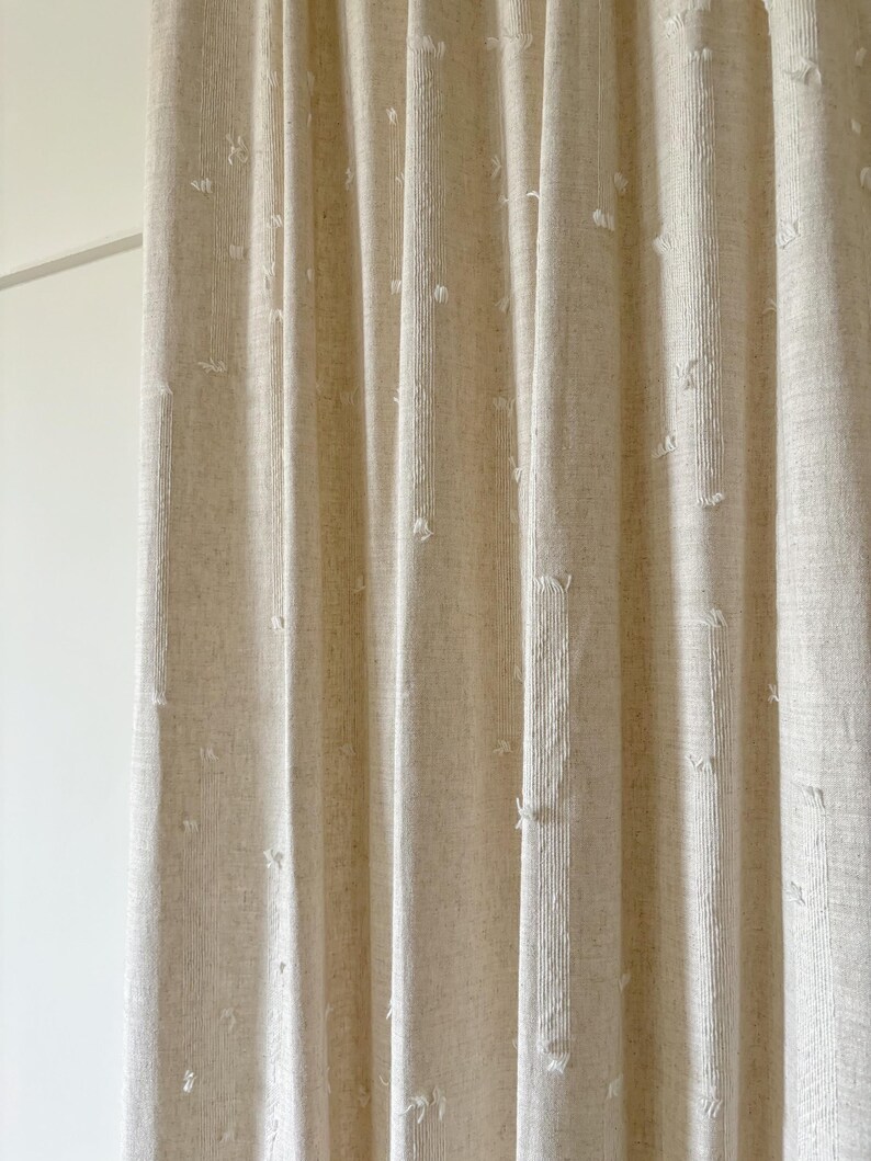 May include: Beige linen curtains with vertical folds. The fabric has a textured appearance with small, irregular white tufts scattered across the surface. The curtains are hanging in front of a white wall.