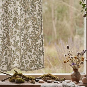 May include: A tab-top curtain featuring a botanical print with green leaves and brown vines on a light beige fabric. The curtain hangs from a rod adorned with a trailing vine, while the window sill displays mossy branches and stones.
