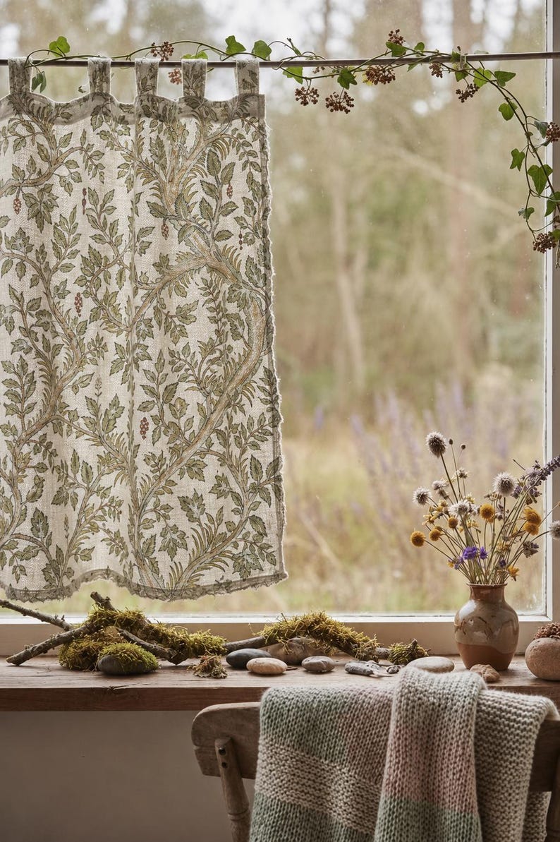 May include: A tab-top curtain featuring a botanical print with green leaves and brown vines on a light beige fabric. The curtain hangs from a rod adorned with a trailing vine, while the window sill displays mossy branches and stones.