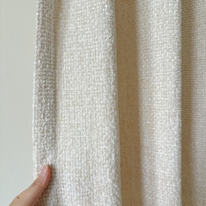 May include: A cream-colored cardigan featuring a textured, woven fabric with a distinct grid-like pattern. The garment has a straight, open-front design with a reinforced edge along the vertical opening, showcasing a soft, slightly fuzzy surface texture.