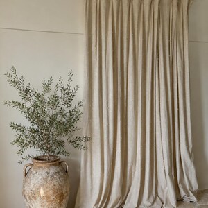 May include: Beige linen curtains hanging from a wooden rod with wooden rings. A large, aged terracotta vase with a green plant sits on a stone floor. The curtains have a textured, dotted pattern.