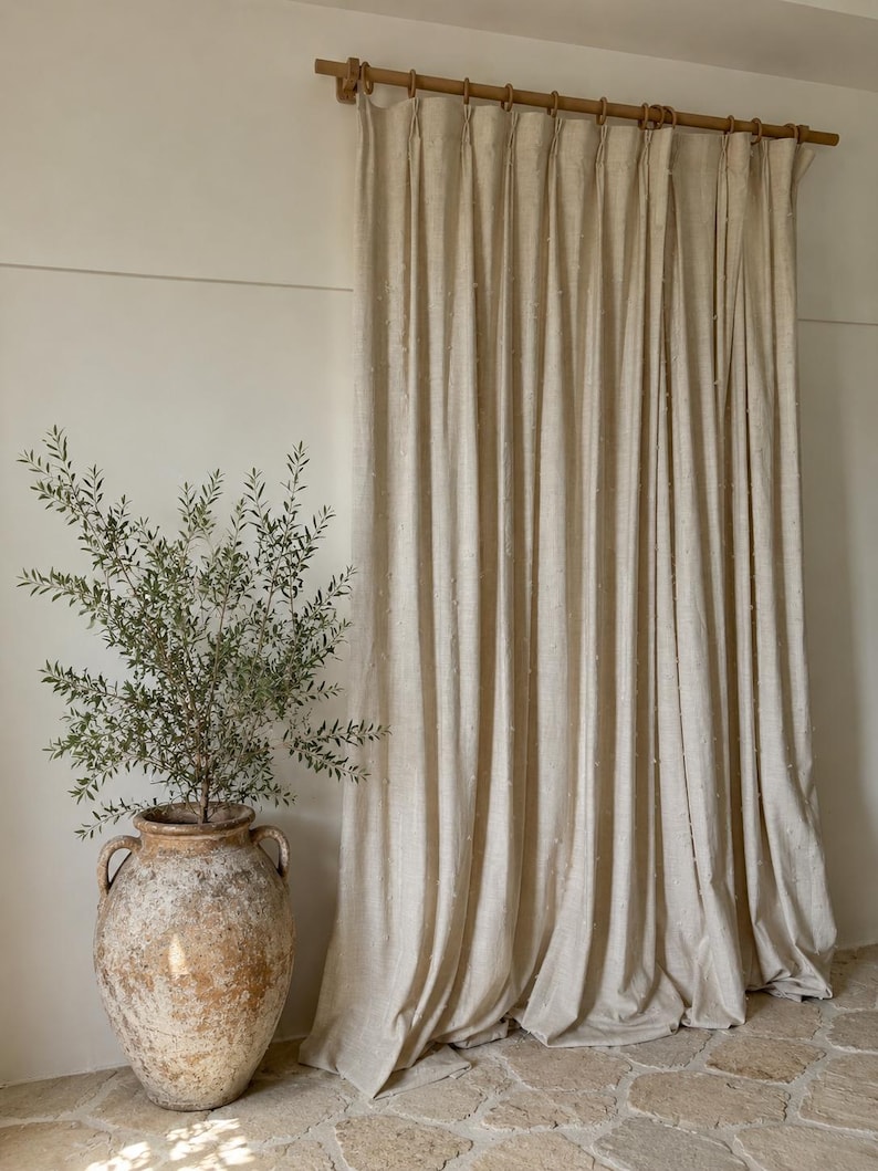May include: Beige linen curtains hanging from a wooden rod with wooden rings. A large, aged terracotta vase with a green plant sits on a stone floor. The curtains have a textured, dotted pattern.