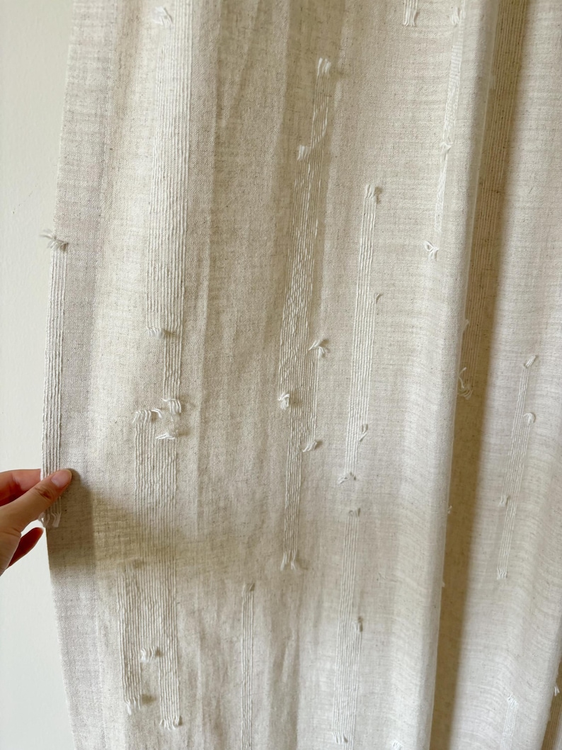 May include: Close-up of a beige linen curtain with vertical stripes and textured details. The curtain has small, raised tufts of fabric scattered across its surface, adding visual interest. The fabric appears to be lightweight and slightly sheer.