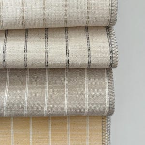 May include: Close-up of four folded fabric samples. The fabrics are various neutral colors, including beige, gray, and yellow, with vertical striped patterns. The edges are finished with a gray stitched border.