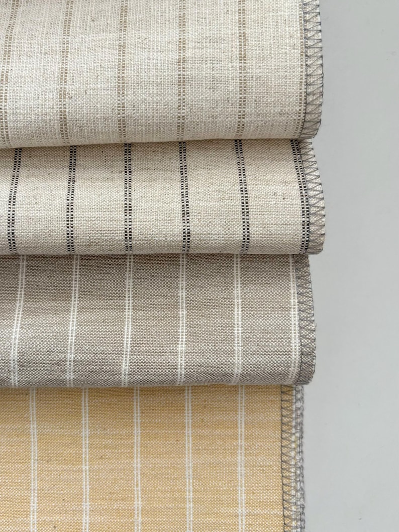 May include: Close-up of four folded fabric samples. The fabrics are various neutral colors, including beige, gray, and yellow, with vertical striped patterns. The edges are finished with a gray stitched border.