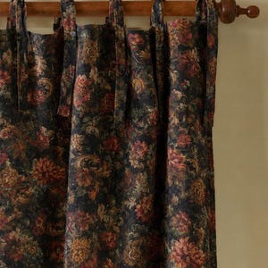 May include: A dark-colored curtain featuring a dense floral pattern in shades of red, orange, and muted green. The curtain is suspended from a wooden rod by several fabric tie-top loops that are knotted at the top of the rod.