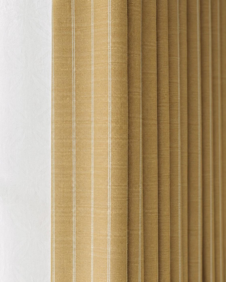 May include: Close-up of a tan fabric with vertical white stripes. The fabric is folded in a pleated style, creating a textured appearance. The color is a warm, neutral tone, suitable for home decor.