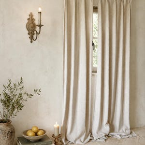 May include: A pair of floor-length, off-white pinch-pleat curtains hanging from a wooden rod with rings. The curtains feature a textured fabric and are positioned over a window, creating a vertical drape that extends to the floor.