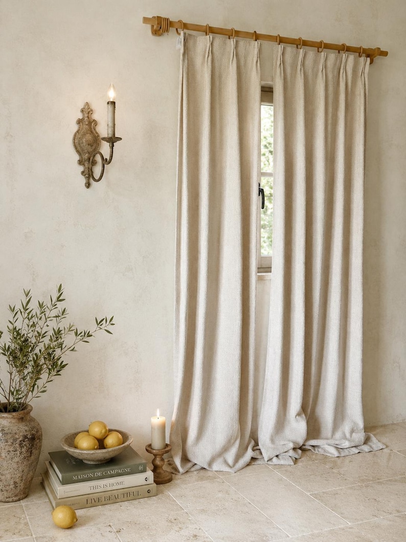 May include: A pair of floor-length, off-white pinch-pleat curtains hanging from a wooden rod with rings. The curtains feature a textured fabric and are positioned over a window, creating a vertical drape that extends to the floor.