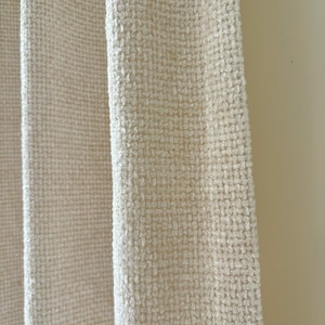 May include: A cream-colored curtain panel featuring a heavy, textured weave with a visible grid-like pattern. The fabric has a thick, slightly raised surface texture that creates depth and dimension across the vertical folds of the material.