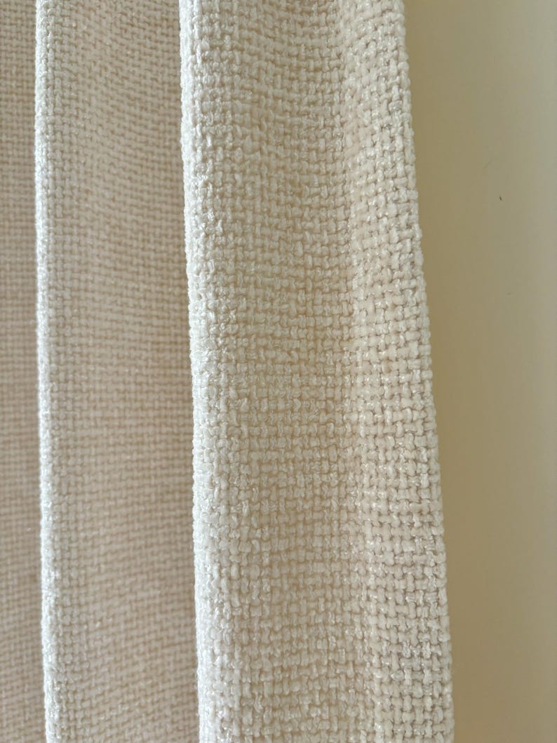 May include: A cream-colored curtain panel featuring a heavy, textured weave with a visible grid-like pattern. The fabric has a thick, slightly raised surface texture that creates depth and dimension across the vertical folds of the material.