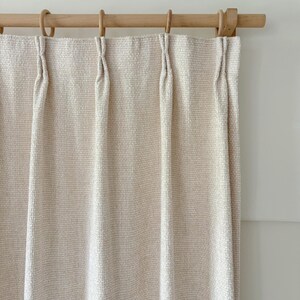 May include: A cream-colored curtain with a textured, woven grid pattern hanging from a light-toned wooden rod. The curtain features evenly spaced pinch pleats at the top, each secured with a circular wooden ring that slides along the rod.