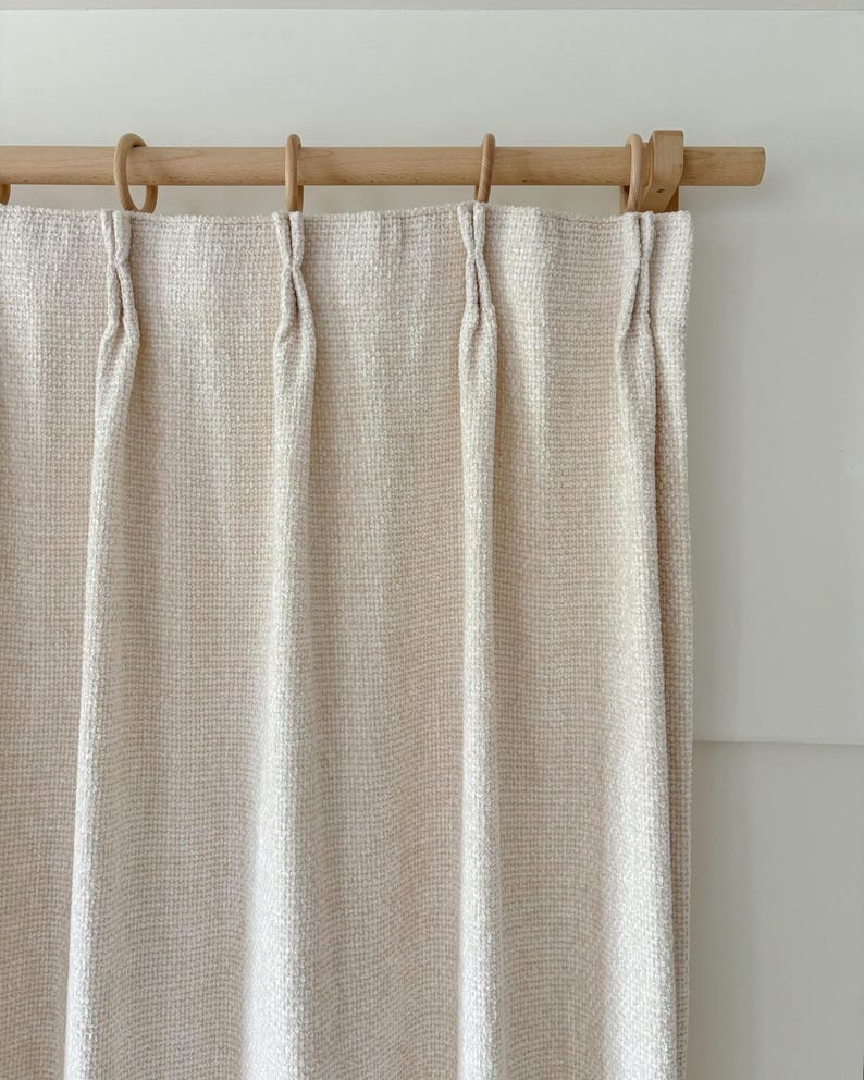 May include: A cream-colored curtain with a textured, woven grid pattern hanging from a light-toned wooden rod. The curtain features evenly spaced pinch pleats at the top, each secured with a circular wooden ring that slides along the rod.
