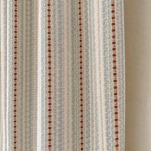 May include: A woven fabric curtain featuring a vertical striped pattern. The design consists of alternating bands of light blue, off-white, and thin, textured rust-colored lines. The material has a visible, tactile weave with a consistent vertical drape.