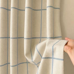 Peut inclure: Un panneau de rideau beige orné d'un motif à carreaux composé de fines lignes verticales et horizontales bleu clair. Le tissu présente un aspect texturé et doux avec des plis verticaux qui créent de la profondeur et du mouvement.