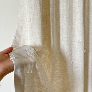 May include: Beige linen curtain with vertical stripes and small, textured embellishments. The fabric is held open, revealing the texture and weave of the material. The curtain appears to be lightweight and semi-sheer, suitable for home decor.