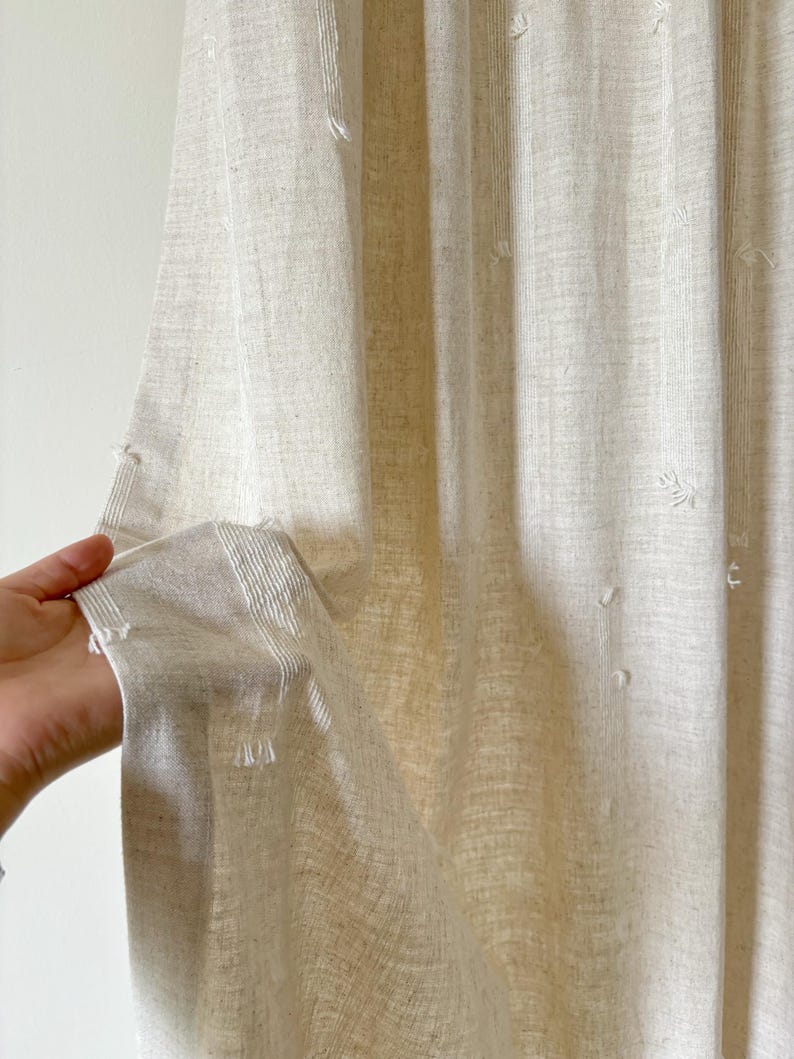 May include: Beige linen curtain with vertical stripes and small, textured embellishments. The fabric is held open, revealing the texture and weave of the material. The curtain appears to be lightweight and semi-sheer, suitable for home decor.