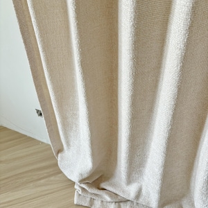 May include: A floor-length, off-white curtain featuring a textured, woven fabric with vertical stripes. The material has a subtle, slightly shimmering finish and hangs in soft, loose folds that extend down to the floor, creating a draped effect.